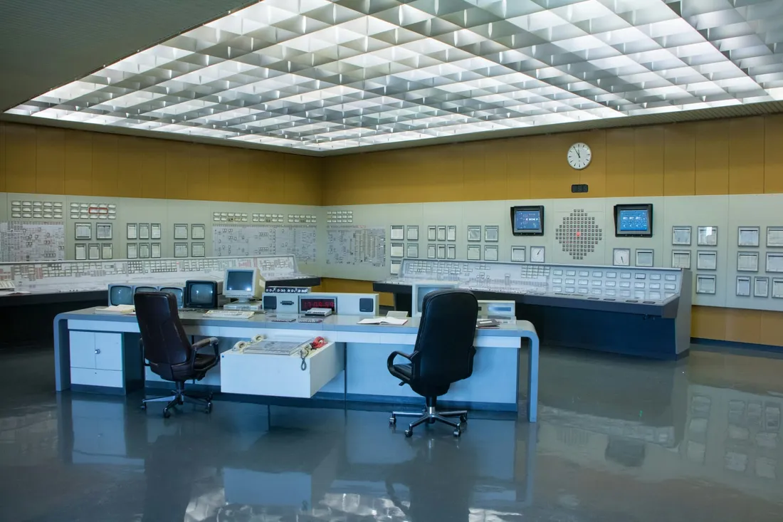 A control room with instrument panels covering the walls from end to end, a clock set among them, and the desks and chairs left empty under a lit ceiling.