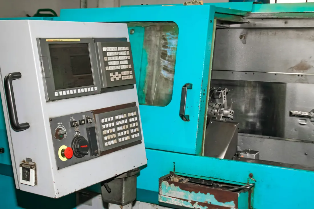 A control console bolted to the side of a machine tool on a shop floor, its screen and keypad set above a red emergency stop, the machine enclosure and its window behind.