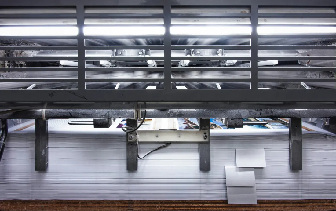 Printed sheets stacked deep under a bank of inspection lamps, one sheet standing proud of the pile and another lying open in the light.